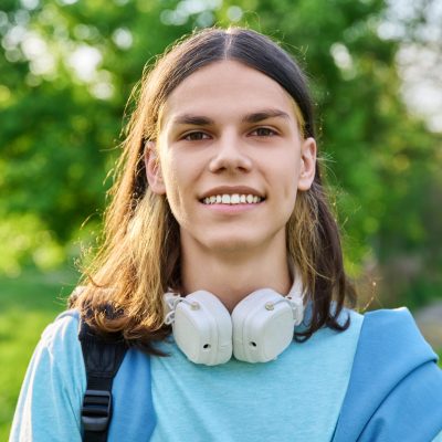 Outdoor portrait of handsome student guy with backpack looking at camera, green park campus background. Young smiling male with long hair hairstyle. Youth, education, young people concept